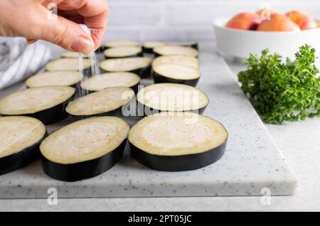 Woman`s hand sprinkles salt over sliced eggplant on a cutting board in the kitchen Stock Photo